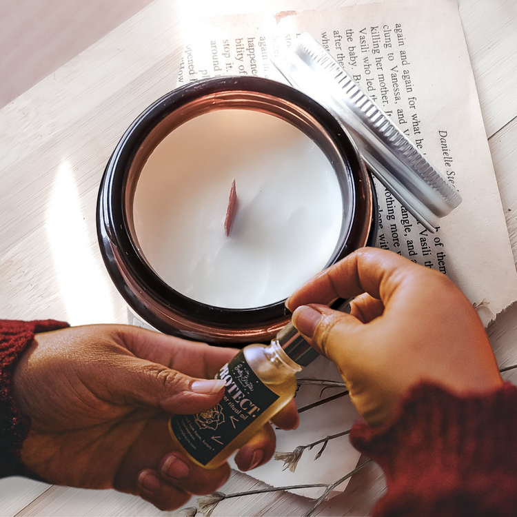 A brown glass candle with white wax over some news paper with two hands holding a frost bottle with a black label and white writing 