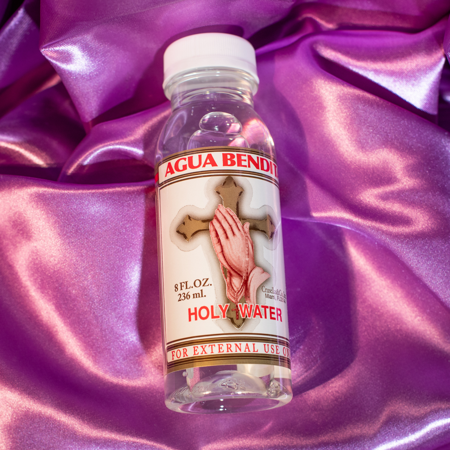 A plastic bottle of 'Agua Bendita' Holy Water by Crusellas & Co., with a label featuring a cross, praying hands and red text on a purple background 