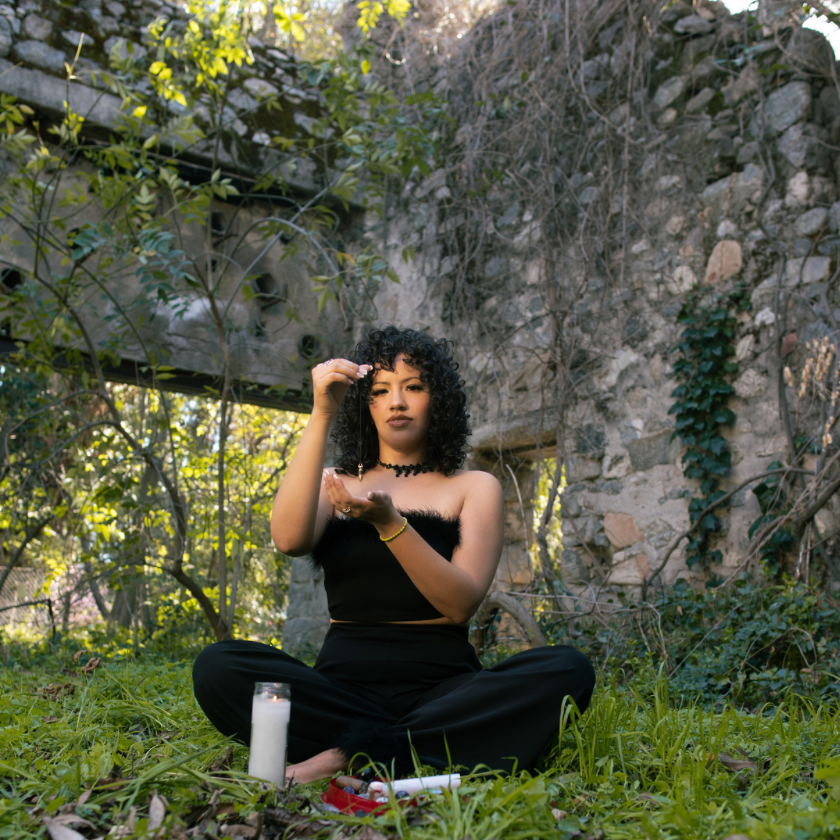 A person sitting cross-legged in a natural setting, with a white candle, holding a pendulum and appearing to be in a state of contemplation or guidance.