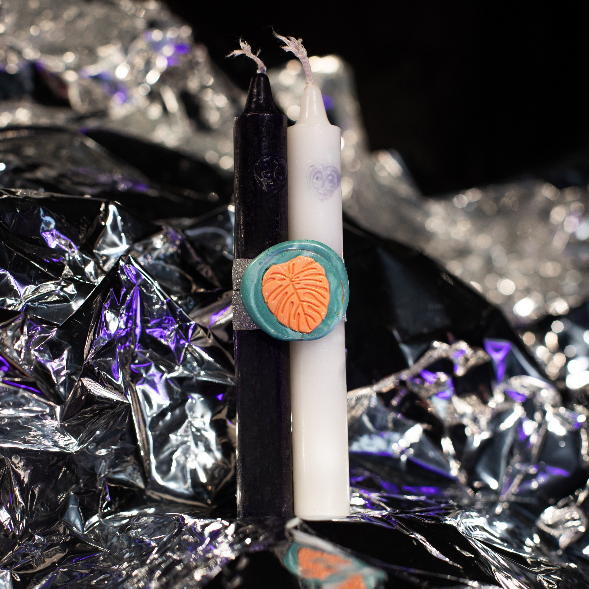 Two candles, one black and one white, placed on a shiny, reflective surface. The candles are held together with an orange and green wax stamp.